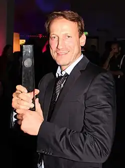 Wotan Wilke Möhring, der beim Deutschen Fernsehpreis 2012 ausgezeichnet wurde. Er trägt einen dunklen Anzug mit Krawatte und hält eine rechteckige, silberne Trophäe in beiden Händen, den Deutschen Fernsehpreis. Der Hintergrund ist dunkel gehalten, mit einigen verschwommenen Lichtquellen und anderen Personen im Hintergrund.