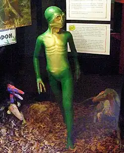 Eine grüne humanoide Gestalt mit gelber Brust deren Gesicht zu einem stumpfen Schnabel zusammenläuft