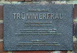 Rechteckige Bronzeplatte mit messingfarbener Aufschrift: Walter Reinhold / Trümmerfrau / 1952 als Eisenguss errichtet / 1967 Neufassung in Bronze / Das Denkmal ist den Frauen Dresdens gewidmet, die in schwerer Zeit mit ihrer Hände Arbeit den Grundstein zum Wiederaufbau der zerstörten Stadt legten / Restaurierung 2001 / Landeshauptstadt Dresden, Stadtsparkasse Dresden, Daimler Chrysler Niederlassung Dresden, Gudrun Benath und Freunde