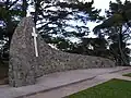 Das Foto eines Denkmals, das vor Bäumen steht. Das Denkmal ist eine Bruchsteinmauer, die einen leichten Bogen beschreibt und auf der linken Seite höher ist als auf der rechten. An der Stelle, an der die Mauer am höchsten ist, befindet sich ein weißes Kreuz auf der Mauer. Am rechten Ende der Mauer ist eine Plakette mit einer Beschriftung zu erkennen. Diese ist allerdings nicht zu lesen. Vor dem Denkmal befindet sich eine Wiese.