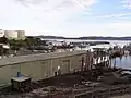 Ein Foto vom Hafen von Eden. In der rechte Bildhälfte ragen einige Stege und eine Kaimauer in ein Gewässer. An den Stegen sind verschiedene etwas ältere Boote befestigt. Im Vordergrund befindet sich ein langgezogener Schuppen aus grünem Wellblech. Hinter diesem befindet sich ein Parkplatz. Vom linken Bildrand ragen große Lagertanks in das Bild. Am anderen Ufer des Gewässers sind Hügelketten zu sehen. Der Himmel ist heiter bis wolkig.