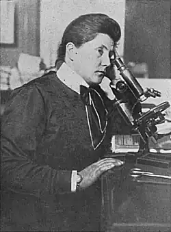 Lydia Kempner-Rabinowitsch sitzt 1912 am Mikroskop, trägt dunkle Kleidung mit hellem Kragen und Halstuch, ihre rechte Hand ruht auf dem Tisch.