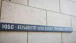 Auf einer Tuffsteinwand ist ein schmaler schwarzer Steinstreifen angebracht, auf dem in heller Schrift „1960 Elisabeth und Eugen Theodor Reintjes“ steht.