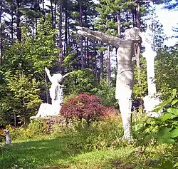 Vier weiße Skulpturen sitzen auf einer mit Gras bewachsenen Lichtung, dazwischen befindet sich ein Busch mit pinkfarbenen Blüten. Im Hintergrund befindet sich ein hochgewachsener Kiefernhain.