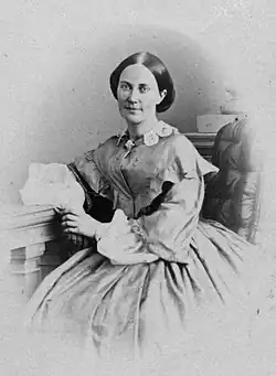 Die Aufnahme der 1860er Jahre zeigt Emma Schenson mit dunklen Haaren, in der Mitte gescheitelt und hinten vermutlich geknotet. Ihr hochgeschlossenes Kleid ist eine Krinoline. Der linke Arm ruht auf einem Gestell mit Säulen.