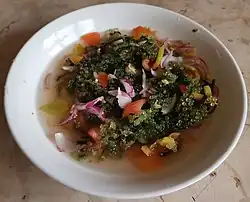 Auf einem weißen Teller ist ein Salat aus Meerestrauben mit in Streifen geschnittenen roten Zwiebeln und Tomatenstücken sowie einem wässrig aussehenden Dressing angerichtet.