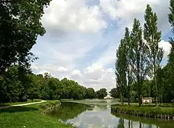 Farbfotografie eines kurvigen Seezufluss, der in der Bildmitte in einen See mit einem hellen Gebäude am hinteren Ufer führt. An beiden Seiten sind Bäume und Wiesen zu sehen, die bis zum hinteren Gebäude führen. Am linken Ufer verläuft ein Weg.