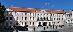 Seitliche Farbfotografie von einem hellroten, dreigeschossigen Gebäude mit zwei Autos auf dem Vorplatz. Der Eingang ist durch einen Vorbau mit Rundbögen erreichbar. Die Fassade ist mit weißen Umrandungen an Fenstern und Ecken verziert. Im Giebelfeld ist ein Wappen und auf dem Dach steht eine blau-schwarz-weiße Flagge.