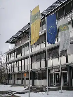 Mehrstöckiges Gebäude von vorn in Frontalansicht mit der mittigen Europaflagge und zwei weiteren Flaggen.