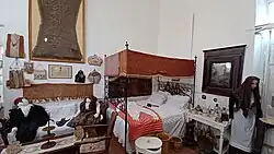 Raum voller historischer Alltagsgegenstände: Bett, Sofa, Tisch, Stühle, Spiegel, drei Puppen in historischer Kleidung