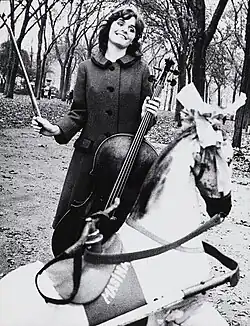 junge Frau mit Cello, in einem Park, vor ihr ein Karussellpferdchen, Schwarzweißaufnahme