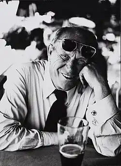 Gert Fröbe sitzt vor einem Bier und lacht in die Kamera, Sonnenbrille auf den Kopf geschoben