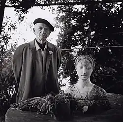 "Otto Dix als älterer Herr, mit Baskenmütze, im Mantel, grimmig schauend, in einem Garten. Rechts im Vordergrund eine antike Frauenbüste