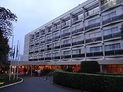Blick von der Hoteleinfahrt auf das fünfstöckige, weiße Gebäude. Die Fenstervorhänge sind zugezogen und vor den Fenstertüren befindet sich jeweils ein sehr schmaler Balkon. Im Erdgeschoss des Hotels brennt Licht und eine Gruppe von Mitarbeitern stehen am überdachten Eingang. Neben der Einfahrt befindet sich eine schmale Grünfläche, die von einer akkurat geschnittenen Hecke eingerahmt ist.