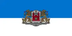Eine blau-weiße Flagge mit einem Wappen in der Mitte. Das Wappen hat eine rote Burg mit einem Löwenkopf in dessen Zentrum sowie eine Krone, ein Kreuz und zwei überkreuzte Schlüssel. Das Wappenschild wird von zwei braunen Löwen gehalten, die ihre Köpfe nach außen gedreht haben und ihre Zunge ausstrecken. Wappen und Löwen stehen auf einem grauen Podest.