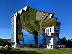 Foto eines ca. 55 Meter breiten und 40 Meter hohen, von Wiese und bewaldeter Landschaft umgebenen Baus, dessen dem Fotografen zugewandte Fassade in voller Breite und Höhe einen riesigen Parabolspiegel bildet. Auf der dem Spiegel abgewandten Seite ist eine von Glas dominierte Vorhangfassade zu erkennen.