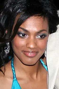 Freema Agyeman mit lächelndem Gesicht, die ein blaues Kleidungsstück mit dünnen Trägern trägt. Ihr dunkles Haar ist in Wellen gestylt, und sie trägt einen auffälligen, glänzenden Ohrring am linken Ohr. Der Hintergrund ist dunkel gehalten, und auf der rechten Seite des Bildes ist ein helles Stoffstück sichtbar, das einen Kontrast zur dunklen Umgebung bildet.