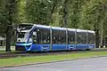 Das Bild zeigt das fünfteilige Fahrzeug von links vorne auf einem breiten Straßenzug mit Grünstreifen und Bäumen im Hintergrund. Der Wagen ist unterhalb der Fenster metallic-blau lackiert, ansonsten schwarz. Die Front ist bullig und mit einer Kühlergrill-Attrappe gestaltet.