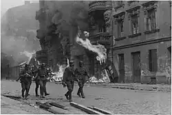 Deutsche Soldaten gehen an Häusern vorbei, aus denen Flammen schlagen; die Straße ist in dichten Rauch gehüllt.