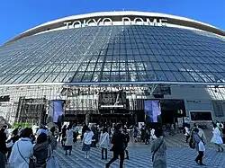 Menschen vor dem Eingang zum Tokyo Dome, unter dem gläsernen Vordach ein Großplakat, das die Show „GIFT“ ankündigt