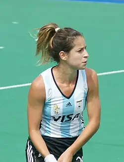 Giselle Kañevsky trägt einen schwarzen Rock und das blau-weiß-gestreifte argentinische Nationaltrikot. Die Haltung der beiden Hände deutet darauf hin, dass sie ihren Schläger führt, dieser ist aber nicht im Bild.