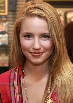 Lächelnde junge Dianna Agron mit langen, blonden Haaren und rot kariertem Hemd. Sie trägt außerdem eine goldene Halskette mit einem großen Anhänger. Der Hintergrund deutet auf einen Innenbereich hin, möglicherweise in einem Geschäft oder Café, da Regale mit verschiedenen Gegenständen sichtbar sind. Die Szene wirkt lebendig und stammt aus der Fernsehserie Glee aus dem Jahre 2009.