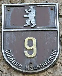 Plakette für Ost-Berlin in Form eines geteilten Wappenschilds, mit dem mauerbekrönten Berliner Bär im ersten Platz sowie montierter goldfarbener Hausnummer im zweiten Platz (Aluminiumguss).