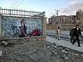 Ein fertiges Graffiti an einer Mauer ist zu sehen. In der rechten Bildhälfte laufen Passanten vorbei und schauen es an.