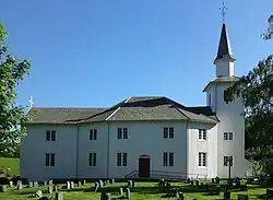 Foto einer weißen Holzkirche. Rechts im Bild der Kirchturm, daran schließt sich ein achteckiger Gebäudeteil an, danach ein Gebäudeteil mit rechteckigem Grundriss. Vor der Kirche befinden sich Grabsteine