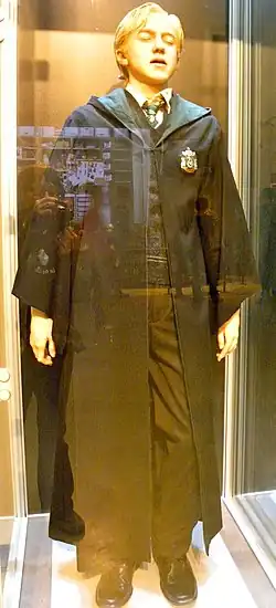 Farbfotografie einer Figur in einer Vitrine, die die Augen geschlossen hat. Sie ist ein Junge und trägt blondes Haar und einen langen schwarzen Umhang mit einem Wappen als Aufnäher.