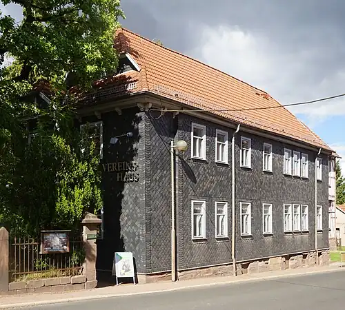 Schrägansicht des ehemaligen Schulhauses. Auf nahezu rechteckigem Grundriss sind Erd- und Obergeschoss vollständig mit Schieferschindeln verkleidet, das Dach ist mit roten Ziegeln gedeckt. Die der Straße zugewandte Traufseite der Fassade hat insgesamt sechzehn hochformatige Fenster. Auf der großteils durch einen Baum verdeckten Giebelseite steht in Metallbuchstaben Vereinshaus.