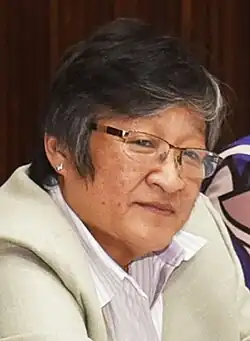 Ein Bild von Helen Mack Chang, der Schwester von Myrna Mack. Es handelt sich um einen Ausschnitt aus einem größeren Bild, bei dem Helen Mack Chang mit weiteren Personen an einem Konferenztisch sitzt. In dem Porträt schaut sie aufmerksam mit leicht gekipptem Kopf in Richtung der Kamera. Sie trägt eine Brille und einen silbernen Ohrstecker sowie ein weißes gestreiftes Hemd und einen hellen Blazer
