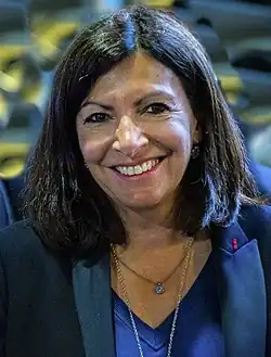 Frontale Farbfotografie einer lächelnden Frau mit dunkelbrauner, schulterlanger Frisur. Sie trägt einen farbigen Anzug und eine dunkelblaue Bluse mit zwei Ketten.