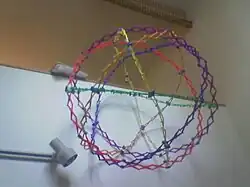 Bunte Kunststoffelemente, die einen Ball aus verschiedenen Kreisflächen formen. Durch Gelenke kann der „Ball“ wachsen und schrumpfen, ohne die allgemeinen Proportionen zu ändern.