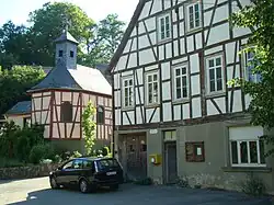 In der rechten Bildhälfte Giebelansicht des früheren Rathauses mit der Schule und einem Schafstall, der sich im massiven Sockelgeschoss des Fachwerkhauses befand. An der Hausfassade befindet sich ein Briefkasten der Post sowie ein Kasten für Bekanntmachungen und Aushänge. An der Hausecke parkt ein Auto. In der linken Bildhälfte etwas im Hintergrund der Chor der Kapelle, ebenfalls ein Fachwerkbau.