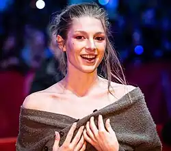 Hunter Schafer steht auf einem roten Teppich und hält ihre Hände mit gespreizten Fingern auf die Brust. Sie trägt einen aufgeknöpften, leicht heruntergezogenen dunkelbraunen Pullover, der ihre Schultern nur teilweise bedeckt, rosa Gelnägel an den Fingern sowie ihre blonden Haare strähnig und hinten über den Rücken, vorne bis zur linken Schulter. Im Hintergrund sind unscharfe Personen, ein dunkelrotes Seitenbanner und dunkelblau-weiße Lichter zu sehen.
