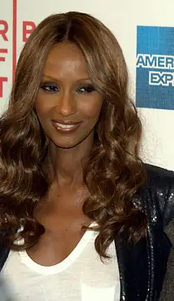 Iman Abdulmajid mit langen, lockigen Haaren und einem eleganten Outfit, lächelt in die Kamera. Sie trägt eine weiße Bluse, die elegant unter einem schwarzen, mit Pailletten besetzten Blazer hervorschaut. Ihr langes, welliges braunes Haar fällt über ihre Schultern. Im Hintergrund ist ein Teil eines Logos zu sehen, das den Text „AMERICAN EXPRESS“ in Blau und Weiß enthält, sowie ein weiteres Element in Rot mit der Aufschrift „R B OF“.