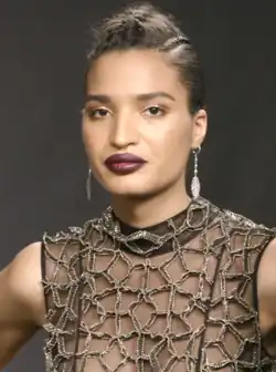 Porträtfoto von Indya Moore. Moore trägt dunkelroten Lippenstift, goldenen Lidschatten, ein dunkelbraunes Top mit einem diamantbesetzten Netzmuster und an beiden Ohren silberne Ohrringe mit an Ketten hängenden Federn. Die braunen Haare trägt Moore als bis zu den Ohren reichende, nach oben abstehende Kurzhaarfrisur.