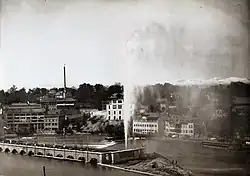 Schwarz-weiss-Bild einer ca. 30 m hohen Fontäne auf einer Insel in der Mitte der Rhone. Das Wasser wird zum Teil vom Winde verweht.