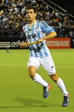 Menini im argentinischen blau-weißen Nationaltrikot mit weißer Hose hält den Hockeyschläger im Lauf in beiden Händen.