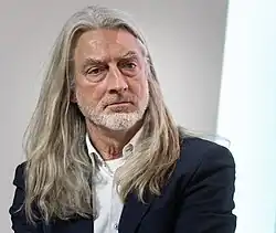 Porträt eines Mannes Anfang 60 mit über schulterlangem grauen Haar, grauem kurzem Bart, weißem Hemd und dunklem Blazer. Blickrichtung schräg nach rechts