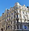 Farbige Eckansicht eines mehrgeschossigen Gebäudes mit runden Eckfassaden. Über und zwischen den Fenstern befinden sich Ornamente. An einigen Fenstern hängen französische Balkone und mehrere Dachgiebeln sowie zwei Fassadentürme sind zu sehen.