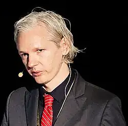 Porträtfoto von Julian Assange mit hellem Teint und schulterlangen, hellblonden Haaren. Er blickt leicht seitlich an der Kamera vorbei und trägt ein dunkles Sakko über einem dunklen Hemd. Der Hintergrund ist unscharf und grau, ohne erkennbare Details.