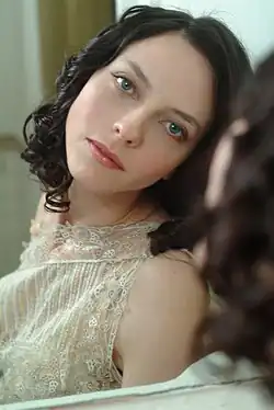 Juliet Landau mit lockigem Haar, die ein Spitzenkleid trägt. Sie schaut verträumt in einen Spiegel, dessen Reflexion im Bild sichtbar ist. Das Kleid ist aus einem filigranen Spitzenstoff mit floralen Mustern gefertigt und scheint hell gefärbt zu sein. Der Hintergrund ist unscharf, sodass keine weiteren Details zu erkennen sind.