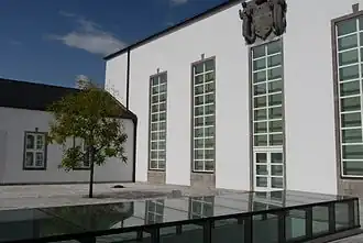Links grüner Baum und kleineres weißes Gebäude, das an ein ebenfalls größes weißes links im rechten Winkel anschließt. Von diesem sind 4 hochgezogene rechteckige Fenster zu sehen. Über das dritte ist ein graues steinernes Wappen platziert. Auf dem gepflasterten Vorplatz ist teilweise ein nach oben hin verglastes niedrigereres Geschoss zu erkennen.