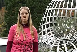 Lemmaperson neben einer rechtsseitigen Skulptur stehend ab Brustbereich als Frontalaufnahme.