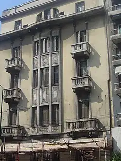 Das Bild zeigt die Frontansicht des 4–5-stöckigen Wohnhauses Papaleonardou. Das Bild zeigt die baufällige Frontansicht bevor es zu einem Hotel renoviert wurde. Das Haus besitzt steinerne Balkone und rechteckige Fenster, ebenfalls mit steinernen Umrandungen.