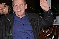 Person Brustbereich in blauem Hemd, schwarzer Jacke von vorn mit gehobenen Arm und in die Kamera lachend, eine weitere lachende männliche Person ist links mit verdrehten Kopf ebenfalls zu sehen.