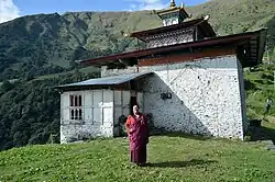 Khochung Lhakhang, Lhuntse, Bhutan. Ein Mönch in Robe vor eine steinernen Gebäude mit weit ausladendem Dach in der Bergkulisse.