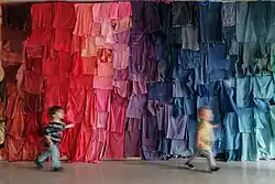 Zwei Kinder laufen im Labor „Glatt oder rau?“ des KinderKunstLabor vor einer regenbogenfarbenen Wand aus Stoffen vorbei.